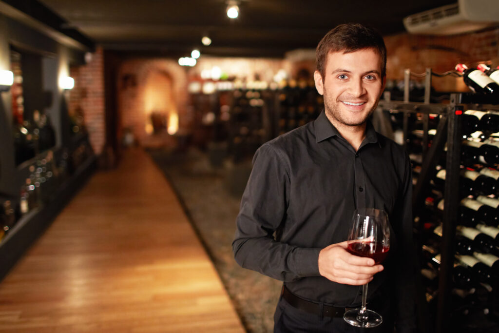 Bild eines jungen Mannes in einem Weinkeller. Er hält ein Glas Wein in der Hand.