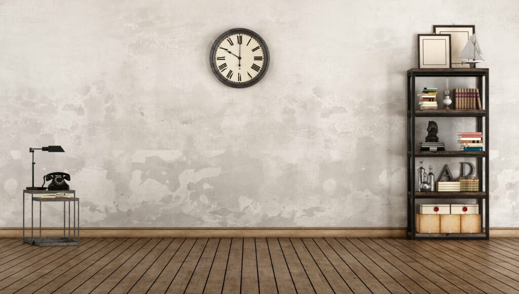 Bild einer grauen Wand und braunem Holzboden. Zu sehen ist eine Ihr, ein mit Büchern und Deko befülltem Regal und ein kleiner Beistelltisch mit Telefon und Lampe.
