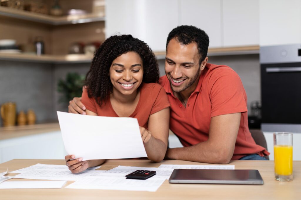 Bild von einer jungen Frau und einem jungen Mann, die lächelnd auf ein Blatt Papier schauen. Vor ihnen liegen weitere Papiere und ein Taschenrechner.