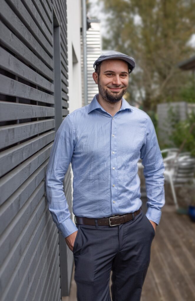 Bild von Malchus Kern. Er trägt eine Anzughose, ein Hemd und ein freundliches Lächeln.