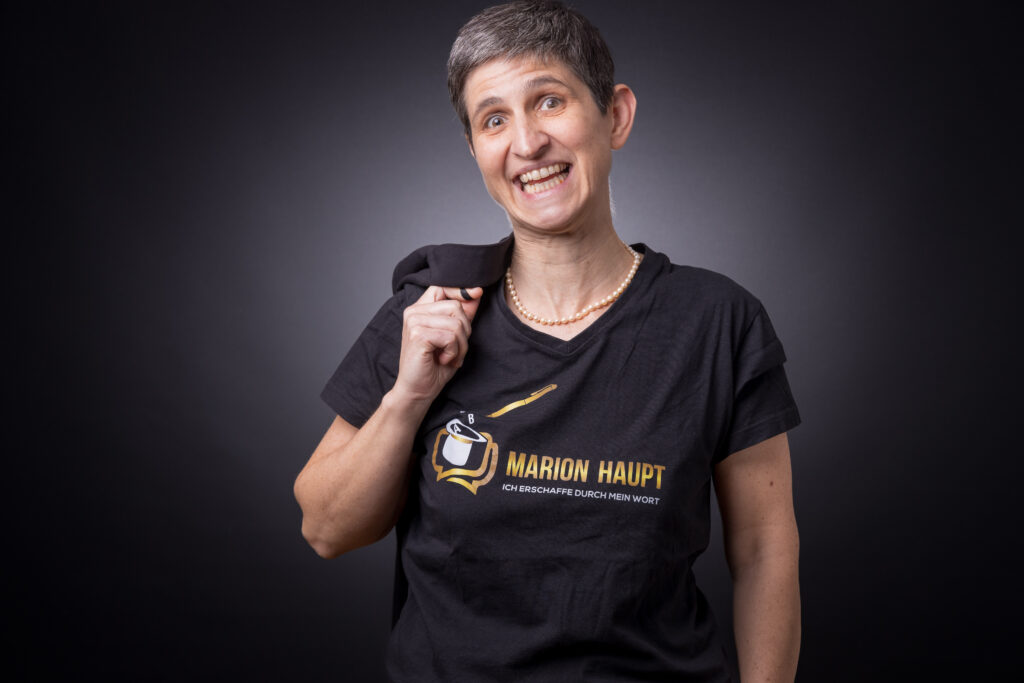Bild von Marion Haupt, sie trägt ein schwarzes T-Shirt mit ihrem Logo und Namen drauf und hat ein weites Lächeln im Gesicht.