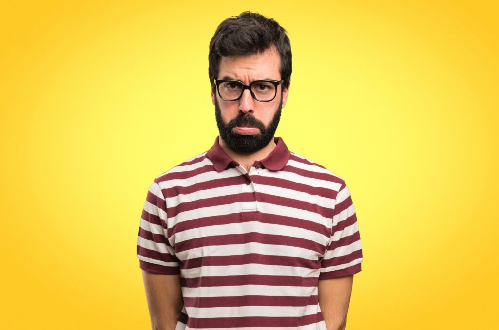 Bild von einem Mann in rot-weiß-gestreiftem T-Shirt vor gelbem Hintergrund. Er trägt eine Brille, hat braunes Haar und zieht einen Schmollmund.