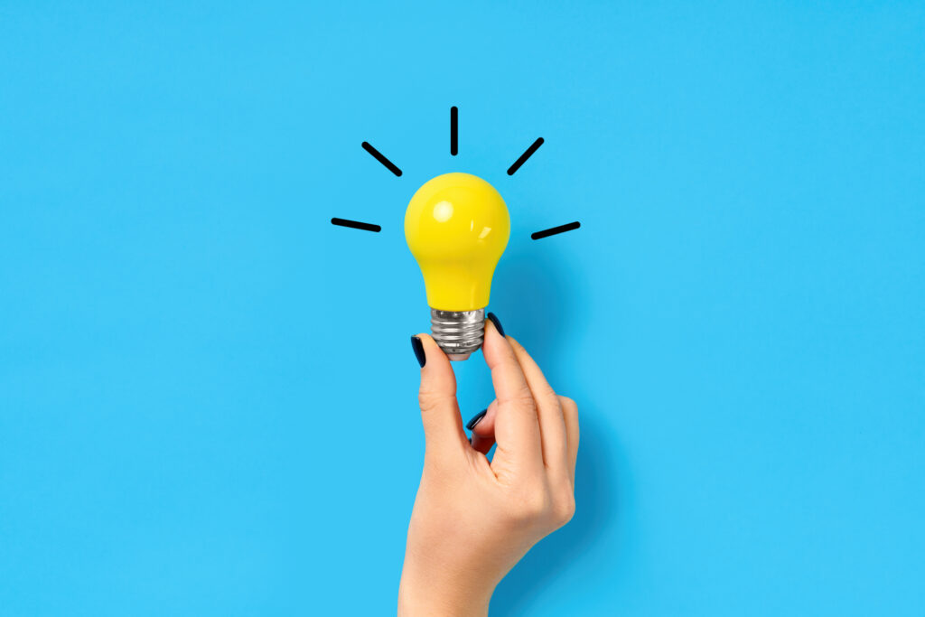 Weibliche Hand hält gelbe Glühbirne vor blauem Hintergrund. Von der Glühbirne abgehende Striche deuten auf Ideen und Brainstorming hin.