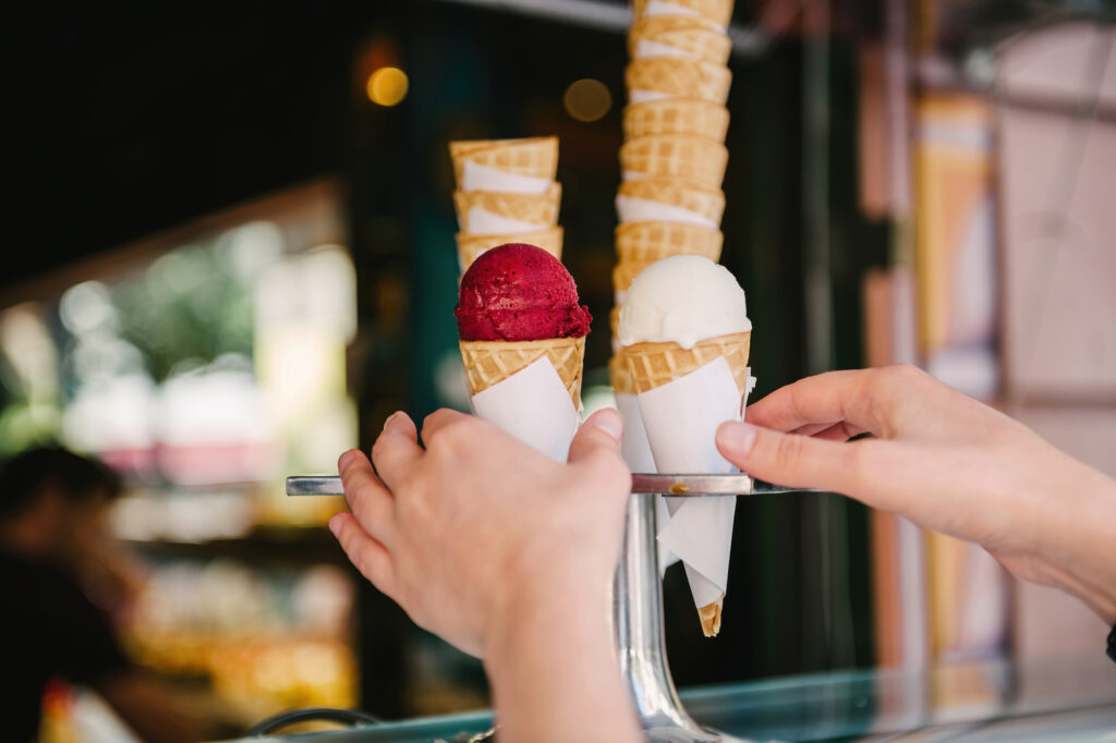 Bild von zwei Händen, die nach zwei Eistüten greifen, in denen jeweils eine rote und eine weiße Kugel Eis drin sind.