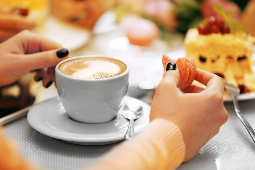 Bild einer Frau, die einen Cappuccino vor sich hat und einen angebissenen Macaron in der Hand hält.